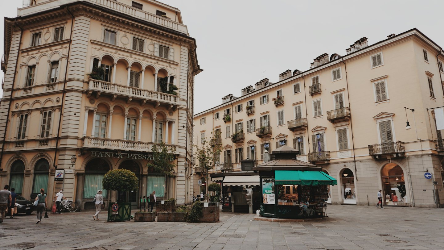 Persone che passeggiano in una piazza cittadina con edifici storici e un'edicola.